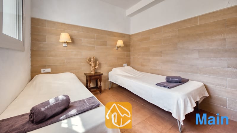Vakantievilla's Costa Brava Spanje - Villa Pilar - Tweepersoons slaapkamer met twee eenpersoonsbedden, opgemaakt met witte lakens en opgevouwen grijze handdoeken, geflankeerd door wandlampen en een klein houten nachtkastje met een decoratieve cactussculptuur.