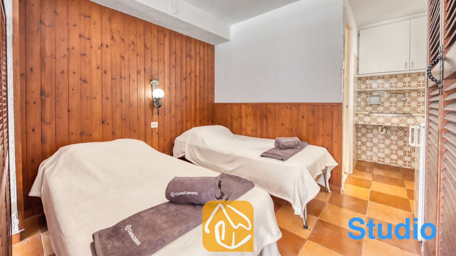 Vakantievilla's Costa Brava Spanje - Villa Pilar - Slaapkamer met twee eenpersoonsbedden voorzien van witte overtrekken en opgevouwen grijze handdoeken, houten wandbekleding en een aangrenzende kitchenette met geprinte tegels bij Villa Pilar.
