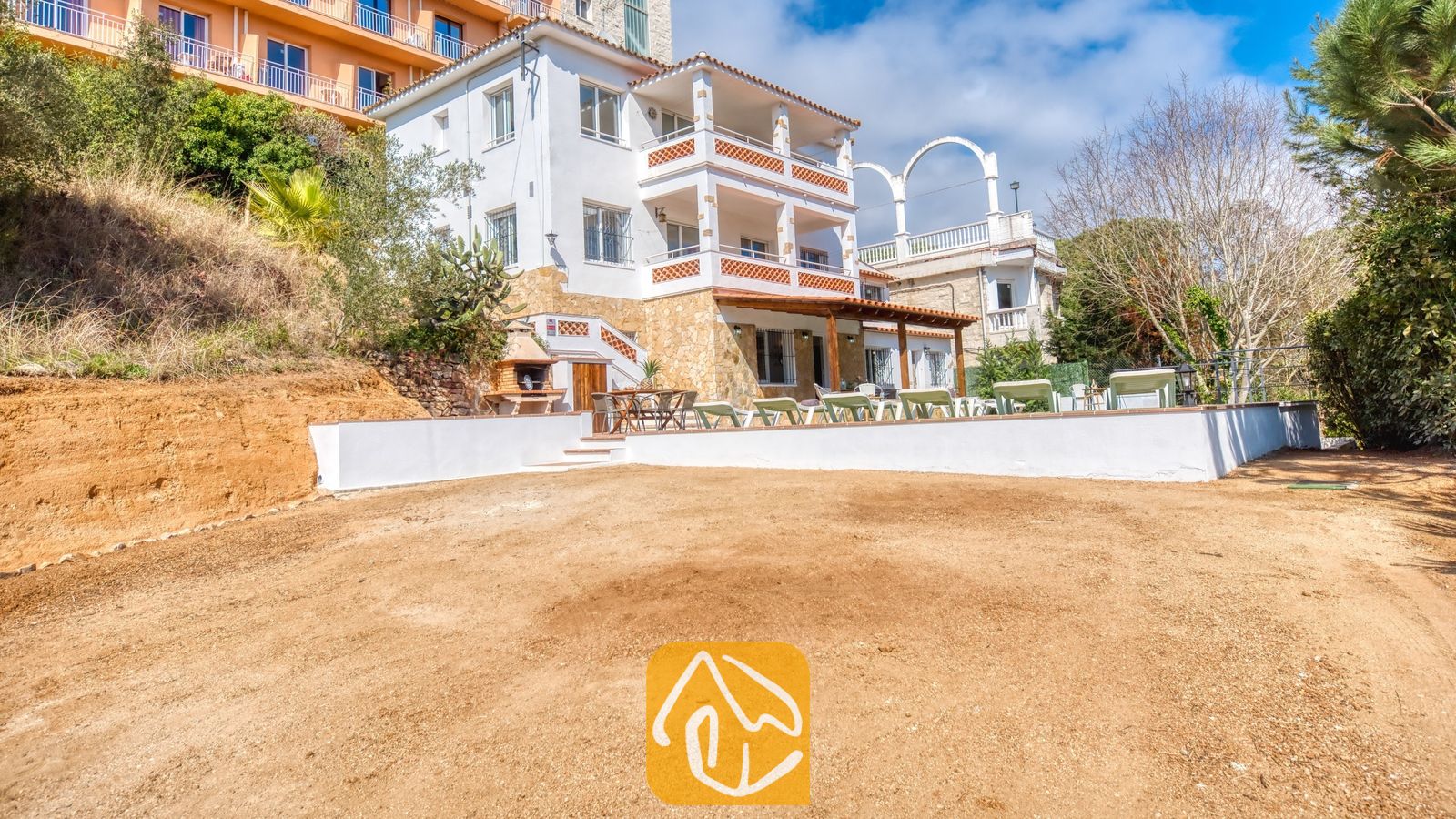 Vakantievilla's Costa Brava Spanje - Villa Pilar - Buitenaanzicht van Villa Pilar met de witte gevel van meerdere verdiepingen, gestapelde balkons met rood getichelde balustrades, stenen sokkelmuren en een onverharde voortuin.
