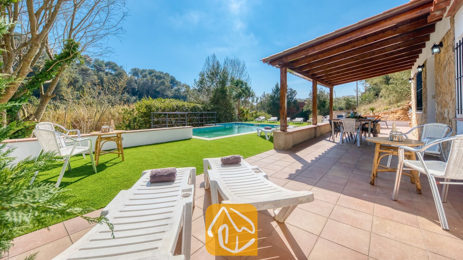 Vakantievilla's Costa Brava Spanje - Villa Pilar - Buitenkant van Villa Pilar met een betegeld terras met ligstoelen, een houten pergola, een eethoek in de buitenlucht en een groen gazon met een zwembad onder een blauwe lucht.