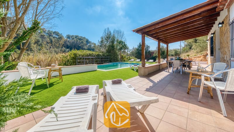 Vakantievilla's Costa Brava Spanje - Villa Pilar - Buitenkant van Villa Pilar met een betegeld terras met ligstoelen, een houten pergola, een eethoek in de buitenlucht en een groen gazon met een zwembad onder een blauwe lucht.
