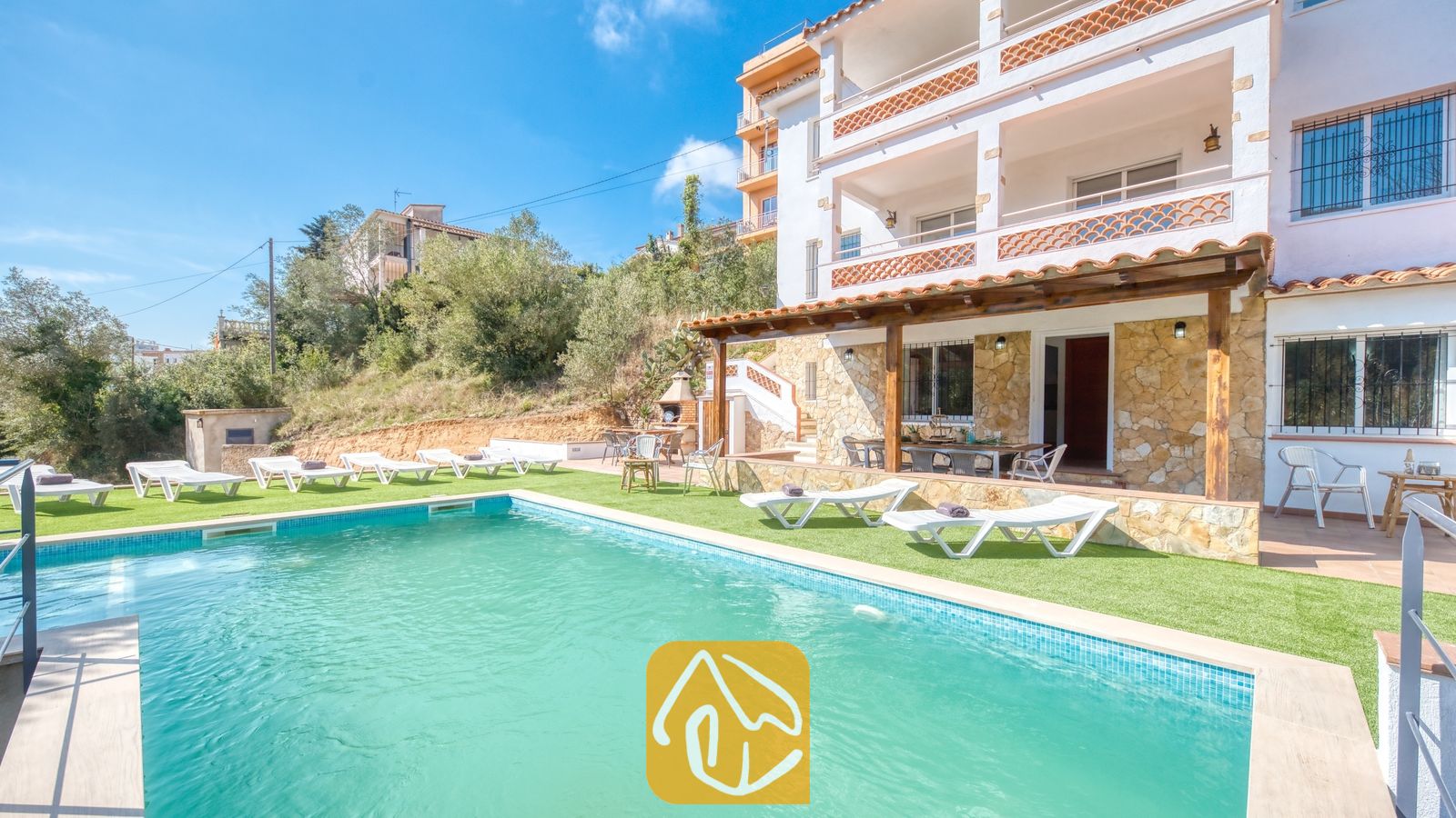 Vakantievilla's Costa Brava Spanje - Villa Pilar - Buitenaanzicht van Villa Pilar met een zwembad, ligstoelen op kunstgras en een wit gebouw met meerdere verdiepingen en stenen accenten.