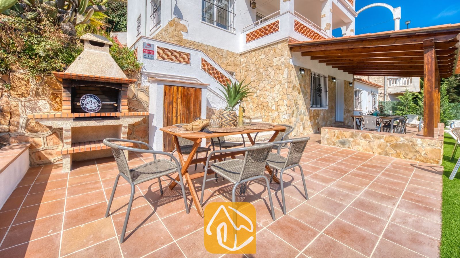 Vakantievilla's Costa Brava Spanje - Villa Pilar - Eethoek buiten bij Villa Pilar met een houten tafel, geweven stoelen en een ingebouwde stenen barbecue op een terras met tegels.