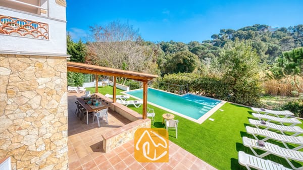 Vakantievilla's Costa Brava Spanje - Villa Pilar - Privézwembad en terras bij Villa Pilar met een houten pergola, een eettafel voor buiten en meerdere witte ligstoelen op kunstgras.