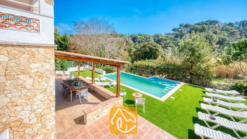 Vakantievilla's Costa Brava Spanje - Villa Pilar - Privézwembad en terras bij Villa Pilar met een houten pergola, een eettafel voor buiten en meerdere witte ligstoelen op kunstgras.