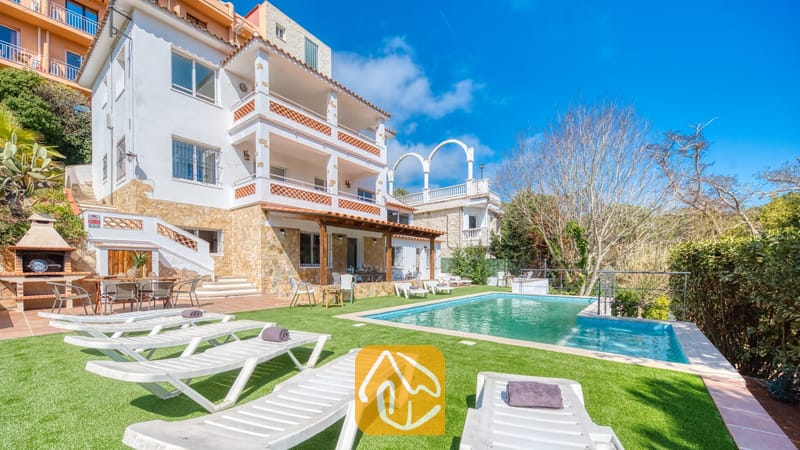 Vakantievilla's Costa Brava Spanje - Villa Pilar - Buitenaanzicht van Villa Pilar met een wit gebouw van meerdere verdiepingen, een stenen sokkel, een zwembad en witte ligstoelen op kunstgras.
