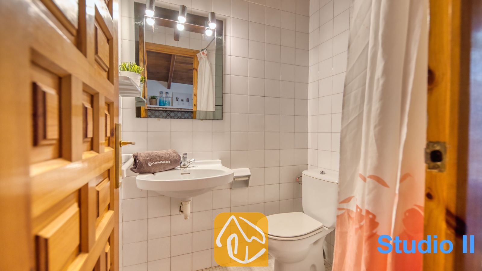 Vakantievilla's Costa Brava Spanje - Villa Pilar - Wit betegeld badkamer met een wastafel aan de muur, een bruine opgevouwen handdoek, een toilet en een verlichte spiegel.