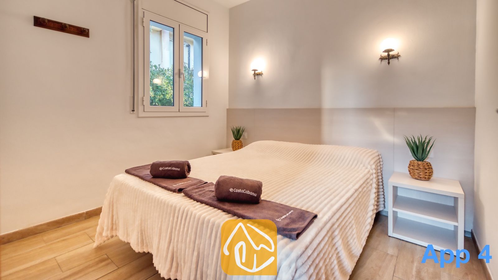 Vakantievilla's Costa Brava Spanje - Villa Pilar - Slaapkamer met een tweepersoonsbed voorzien van een gestructureerde witte sprei, opgerolde bruine handdoeken en twee nachtkastjes met in pot geplante planten onder wandlampen.