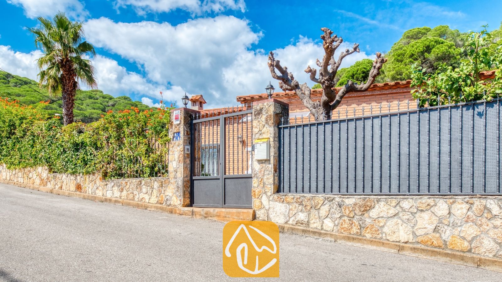 Vakantievilla's Costa Brava Spanje - Villa Alba - De buitengevel en metalen toegangspoort van Villa Alba, met stenen muren, een hoge palmboom en een geplaveide straat tegen een blauwe hemel.