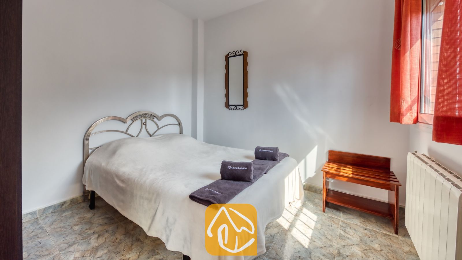 Vakantievilla's Costa Brava Spanje - Villa Alba - Slaapkamer met een wit bed, grijs gevouwen handdoeken, een metalen hoofdbord, rode gordijnen en een houten bagagerek.