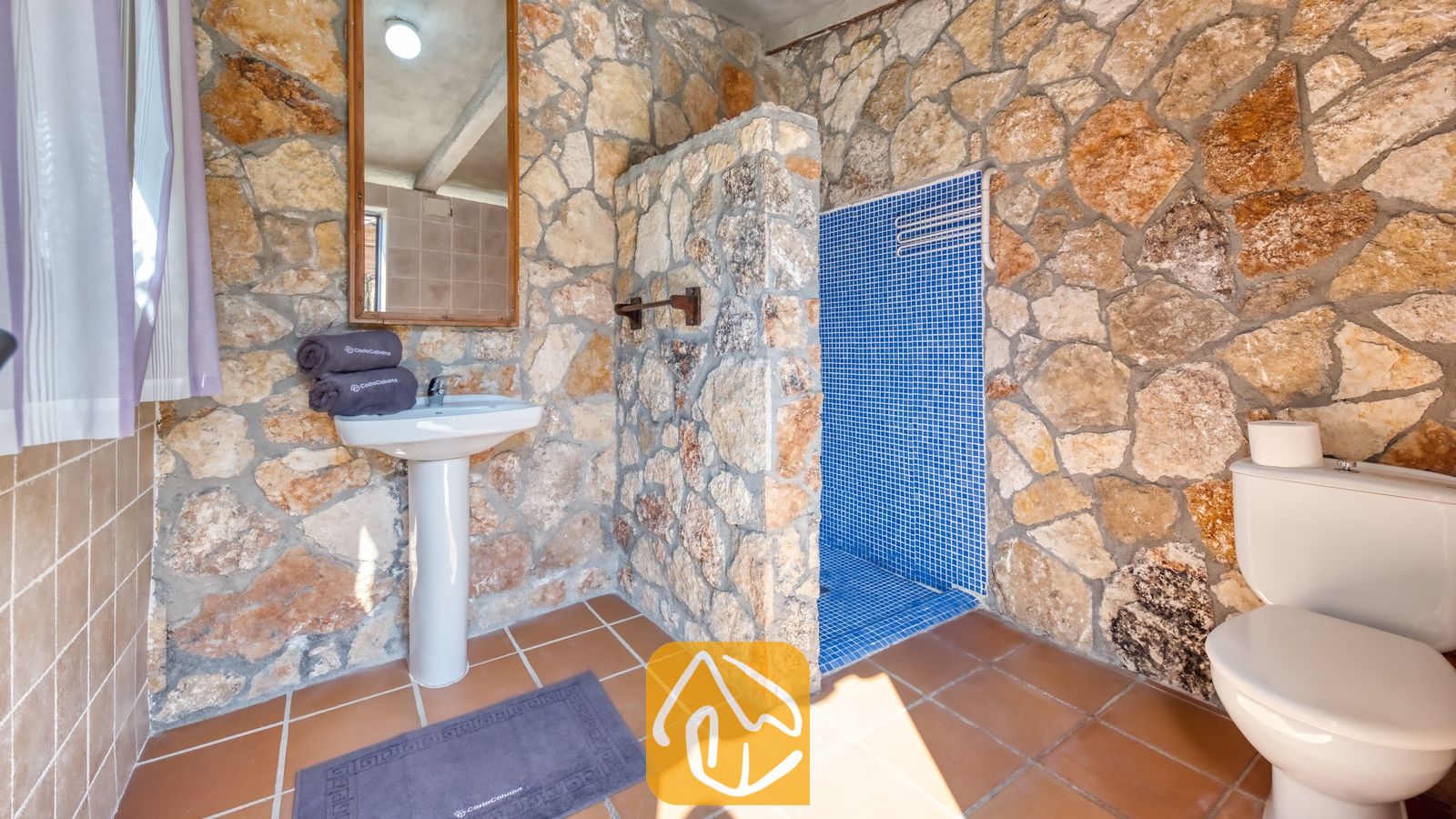 Vakantievilla's Costa Brava Spanje - Villa Alba - Badkamer met rustieke stenen muren, een witte zuilwastafel, een betegelde doucheruimte met blauwe mozaïeken en bruine vloertegels.