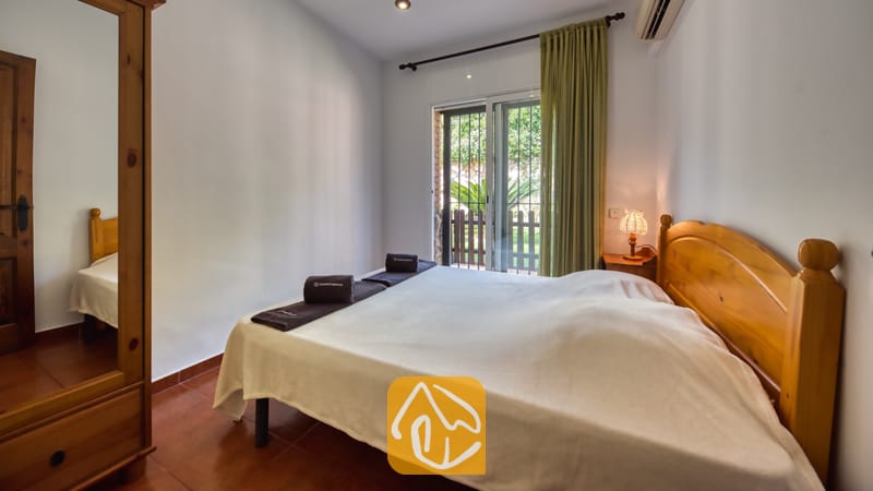 Vakantievilla's Costa Brava Spanje - Villa Alba - Slaapkamer met een houten tweepersoonsbed, bijpassende kledingkast met spiegel, opgerolde donkere handdoeken en een glazen schuifpui die toegang biedt tot een balkon.