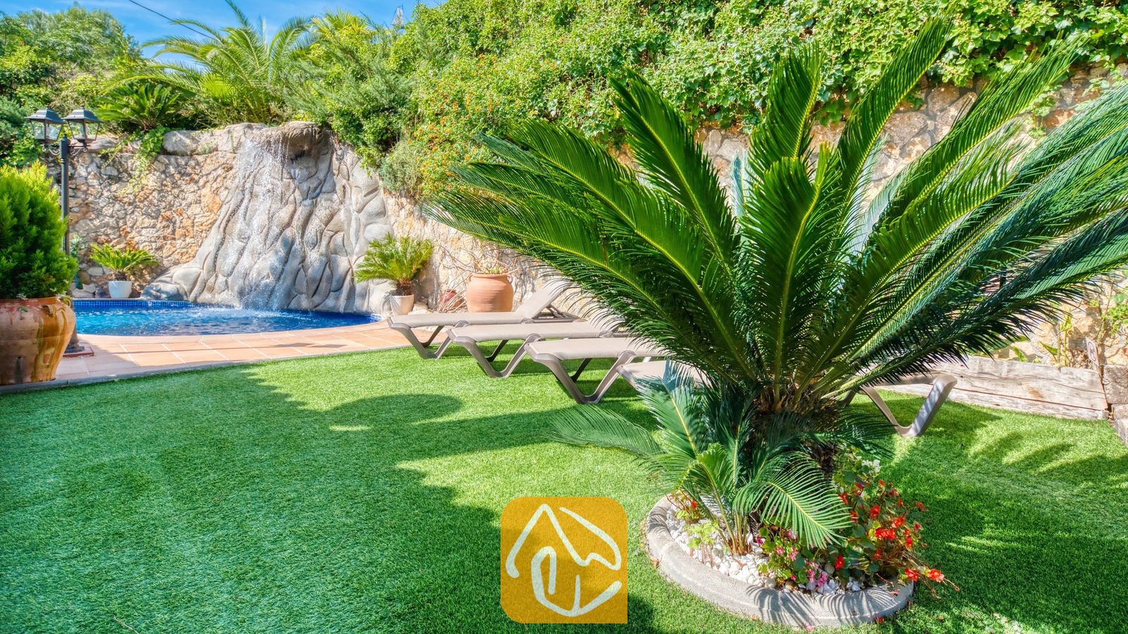 Vakantievilla's Costa Brava Spanje - Villa Alba - Weelderige groene tuin bij Villa Alba met kunstgras, een opvallende cycadale palm, ligstoelen en een stenen waterpartij met waterval.