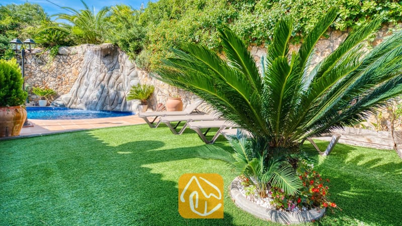 Vakantievilla's Costa Brava Spanje - Villa Alba - Weelderige groene tuin bij Villa Alba met kunstgras, een opvallende cycadale palm, ligstoelen en een stenen waterpartij met waterval.