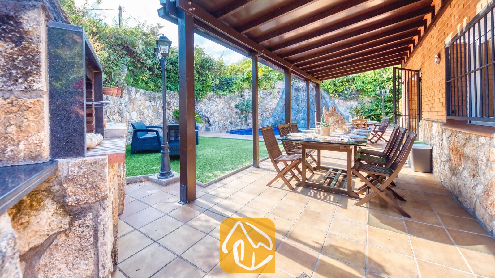 Vakantievilla's Costa Brava Spanje - Villa Alba - Overdekt buitenterras bij Villa Alba, voorzien van een houten eettafel met stoelen, een tegelvloer en een aangrenzende, ingebouwde barbecuehoek met uitzicht op de tuin en het zwembad.