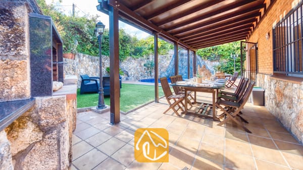 Vakantievilla's Costa Brava Spanje - Villa Alba - Overdekt buitenterras bij Villa Alba, voorzien van een houten eettafel met stoelen, een tegelvloer en een aangrenzende, ingebouwde barbecuehoek met uitzicht op de tuin en het zwembad.