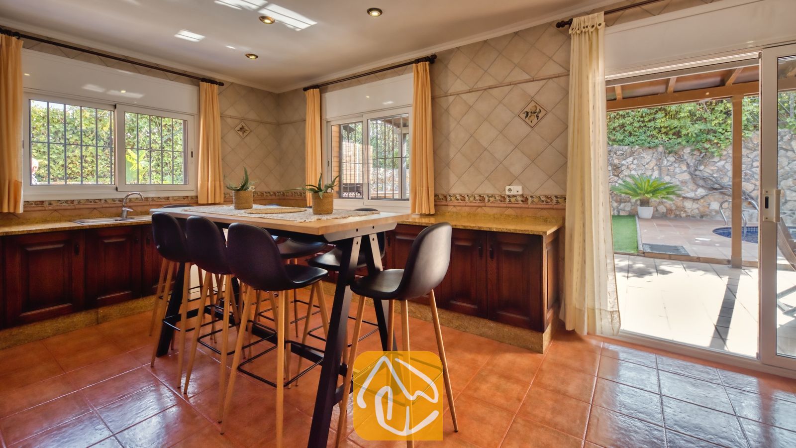 Vakantievilla's Costa Brava Spanje - Villa Alba - Keuken met eethoek voorzien van een hoge houten tafel en zwarte barsturelen, terracotta vloertegels, betegelde muren en een open glazen deur die leidt naar een buitenterras bij Villa Alba.