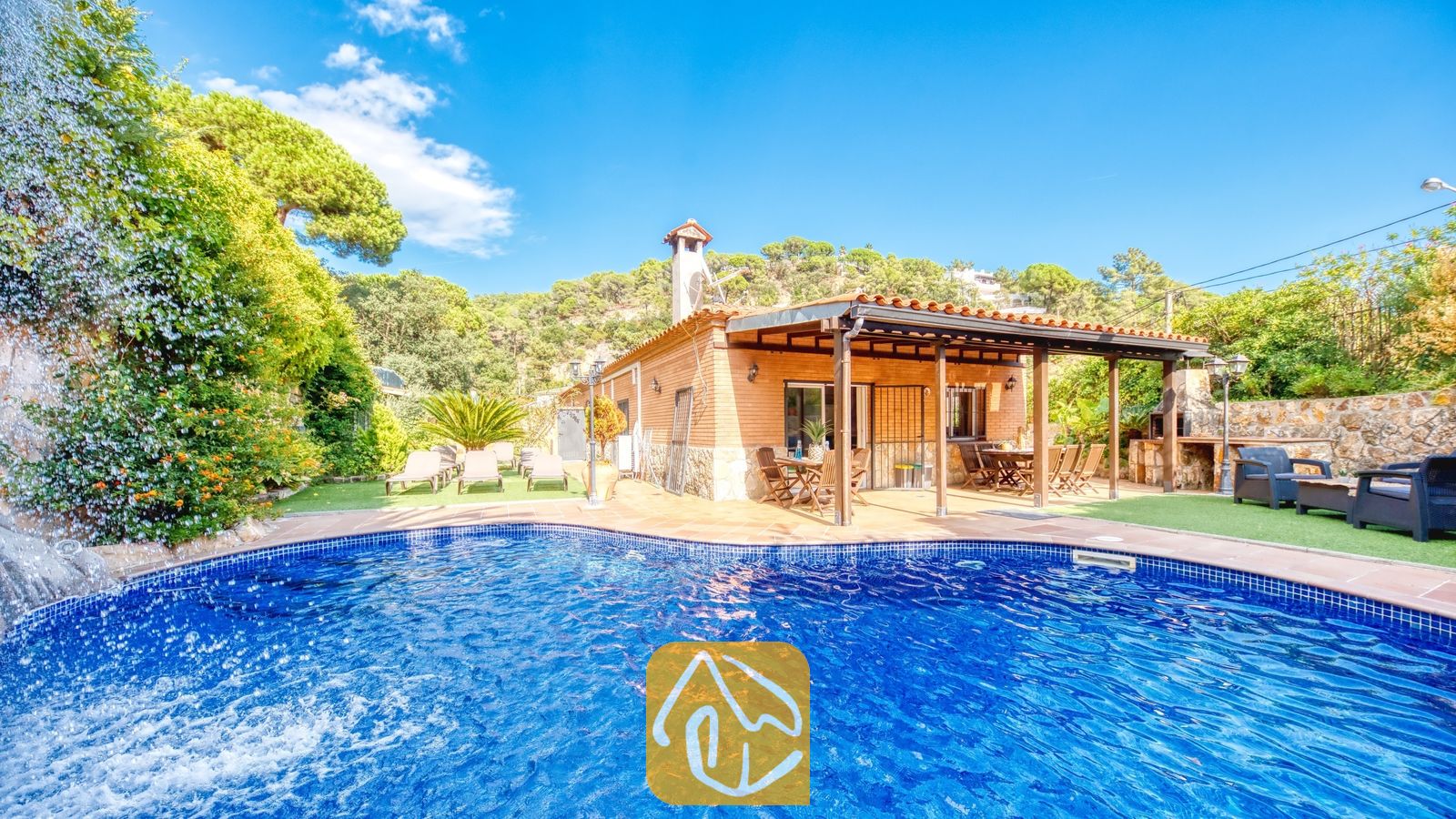 Vakantievilla's Costa Brava Spanje - Villa Alba - Privézwembad met een stenen waterval bij Villa Alba, met een bakstenen gevel, een overdekt terras en ligstoelen op kunstgras onder een blauwe hemel.