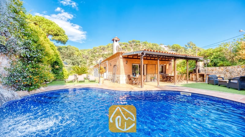 Vakantievilla's Costa Brava Spanje - Villa Alba - Privézwembad met een stenen waterval bij Villa Alba, met een bakstenen gevel, een overdekt terras en ligstoelen op kunstgras onder een blauwe hemel.