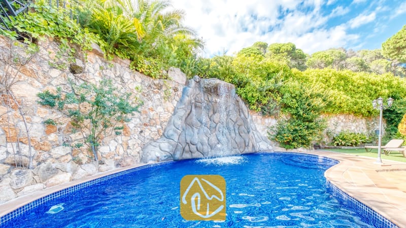 Vakantievilla's Costa Brava Spanje - Villa Alba - Een rotswaterval die uitmondt in een blauw getegeld zwembad bij Villa Alba, omringd door stenen keermuren en groen loof onder een licht bewolkte hemel.