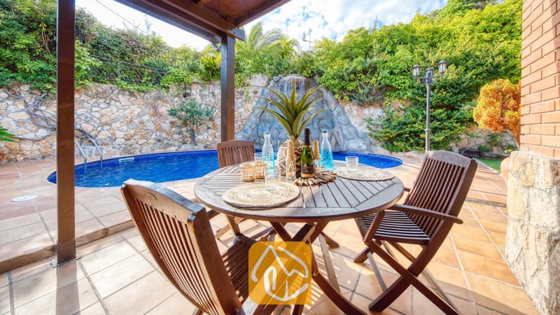 Vakantievilla's Costa Brava Spanje - Villa Alba - Houten eettafel en stoelen op een stenen terras bij Villa Alba, met een blauw zwembad en een rotswaterval als blikvanger op de achtergrond.
