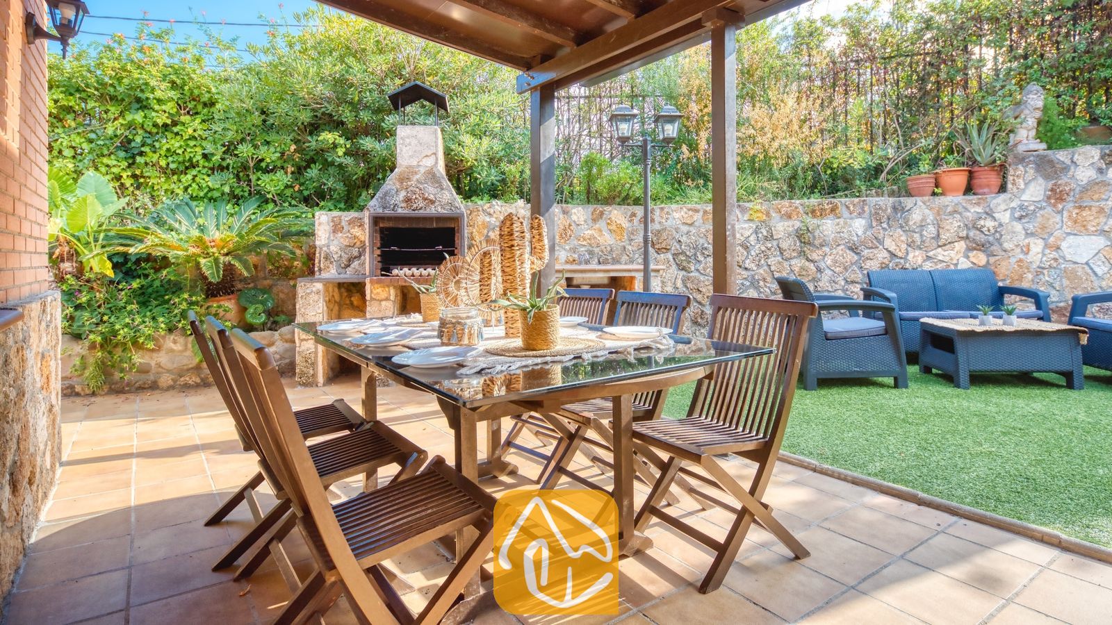 Vakantievilla's Costa Brava Spanje - Villa Alba - Overdekt buitenbeterterras bij Villa Alba met een houten tafel met glazen blad, klapstoelen en een ingebouwde stenen barbecue op de achtergrond.