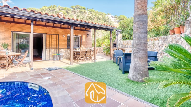 Vakantievilla's Costa Brava Spanje - Villa Alba - Buitenvoorzieningen en zwembadzone bij Villa Alba, met een overdekt terras met houten balken, een eethoek buiten en een gazon omzoomd door een stenen muur