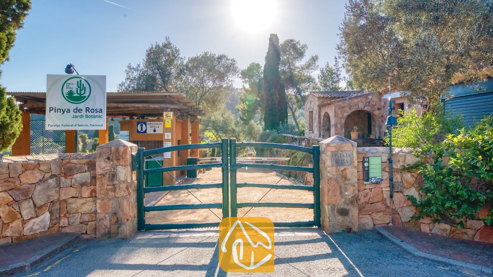 Vakantievilla's Costa Brava Spanje - Villa Santa Cristina - Groene metalen toegangspoort tot de botanische tuin Pinya de Rosa nabij Villa Santa Cristina, gekenmerkt door stenen muren, informatieborden en hoge cipressen in het felle zonlicht.