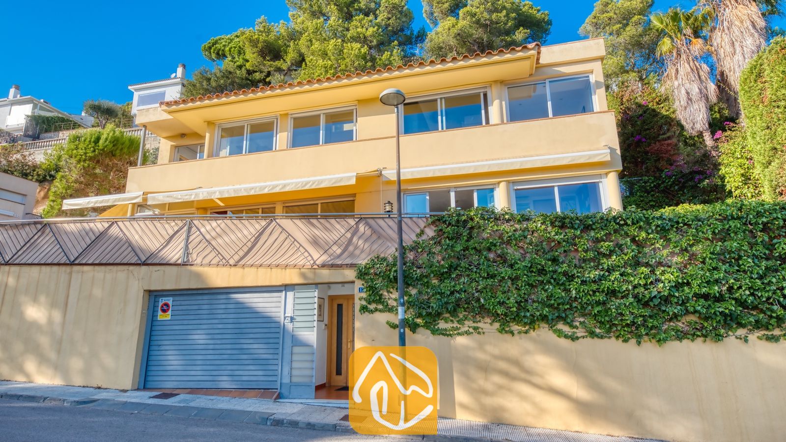 Vakantievilla's Costa Brava Spanje - Villa Santa Cristina - De geelgekleurde, meerlaagse gevel van Villa Santa Cristina valt op door de grote glazen raampartijen, witte zonneschermen en een grijze garagedeur. De woning wordt omringd door weelderige klimop en statige dennenbomen.