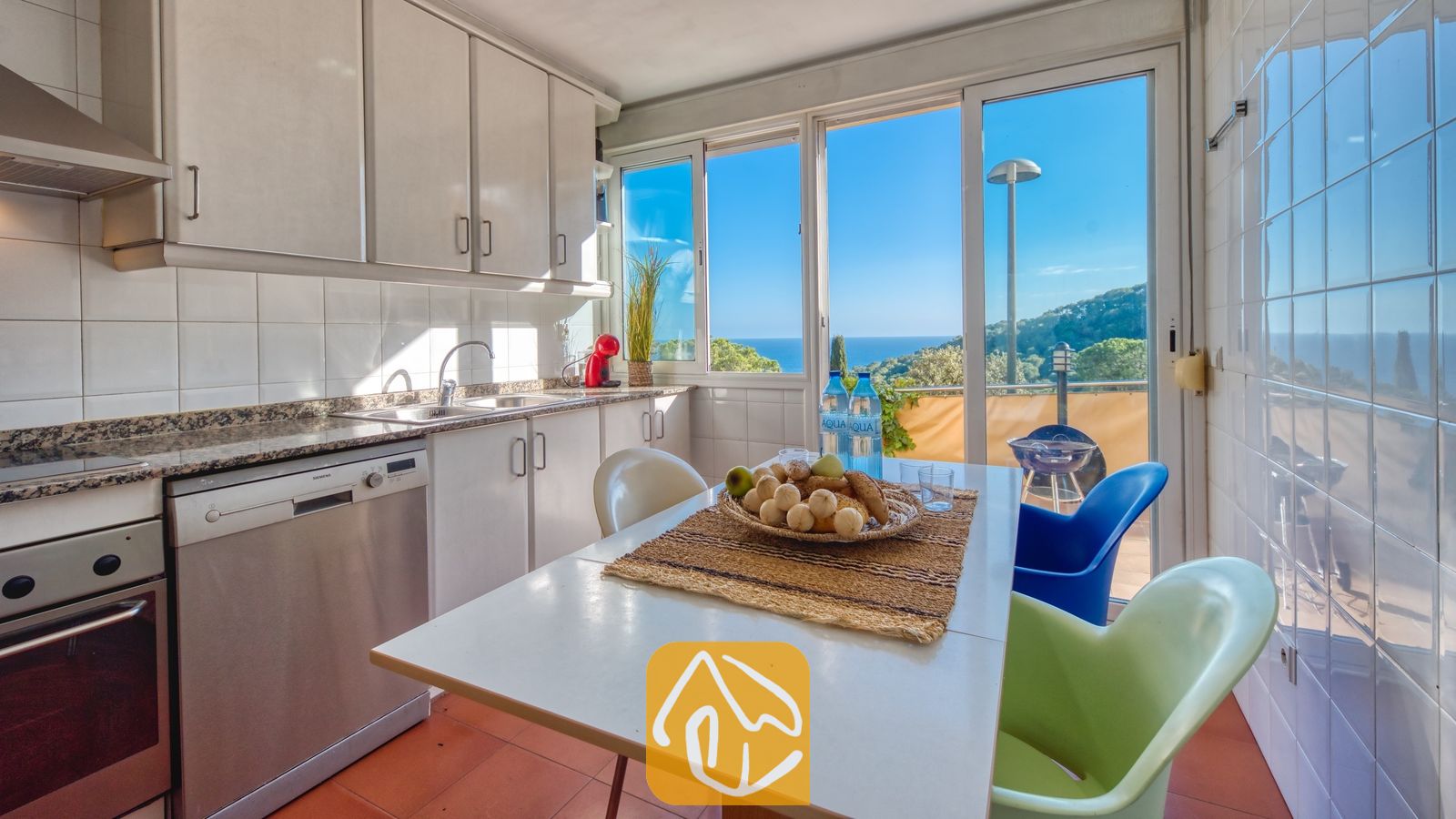 Vakantievilla's Costa Brava Spanje - Villa Santa Cristina - De keuken van Villa Santa Cristina is voorzien van witte kasten, een eettafel met kleurrijke stoelen en een groot raam met uitzicht op de zee en de groene heuvels.