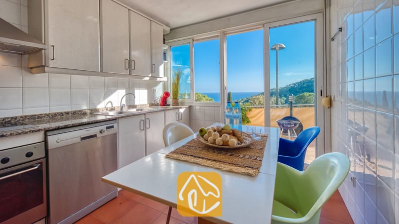 Vakantievilla's Costa Brava Spanje - Villa Santa Cristina - De keuken van Villa Santa Cristina is voorzien van witte kasten, een eettafel met kleurrijke stoelen en een groot raam met uitzicht op de zee en de groene heuvels.