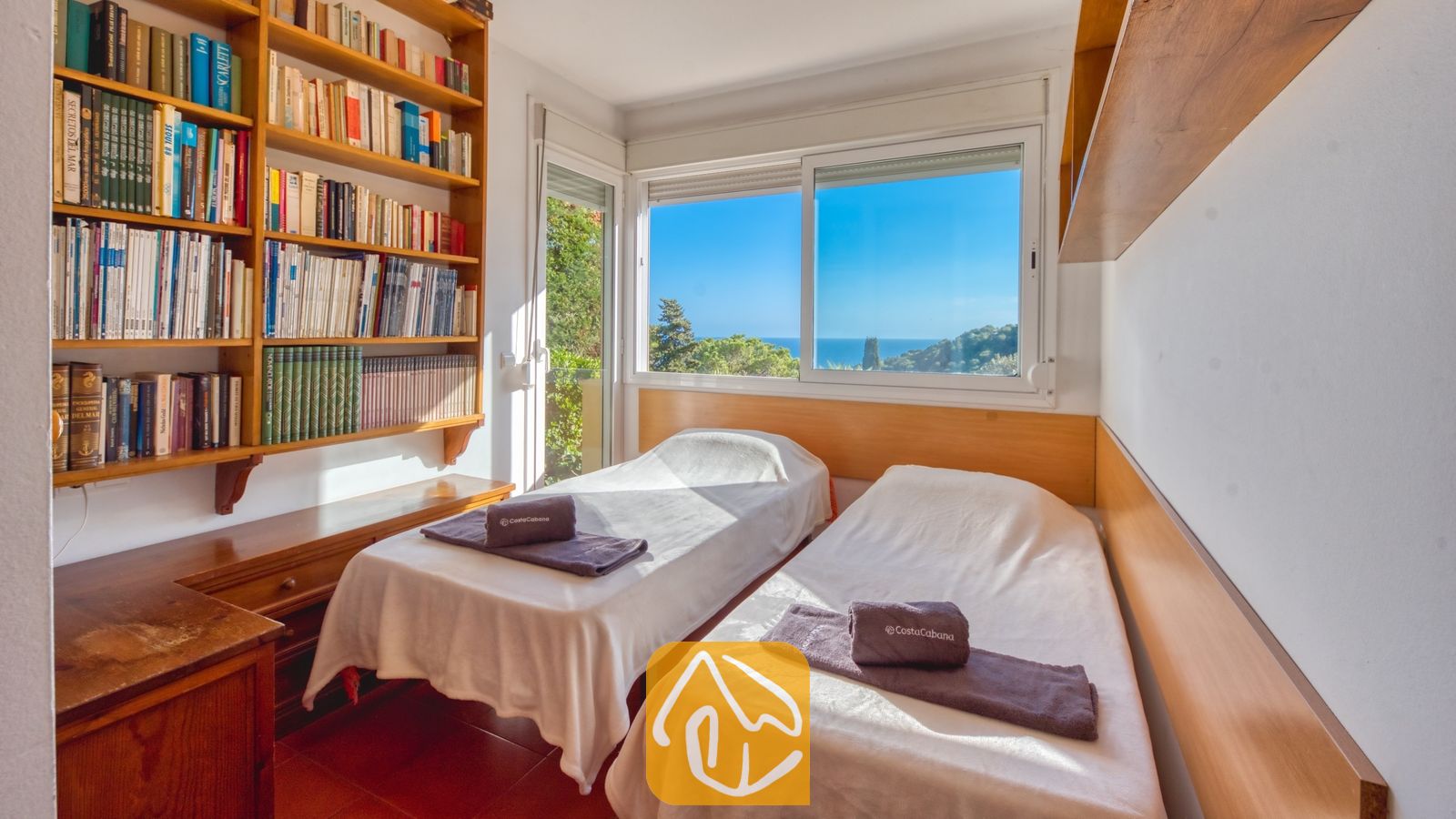 Vakantievilla's Costa Brava Spanje - Villa Santa Cristina - Slaapkamer in Villa Santa Cristina met twee eenpersoonsbedden, wit beddengoed en grijze handdoeken. Naast het raam, dat een prachtig uitzicht op zee biedt, staat een grote houten boekenkast.