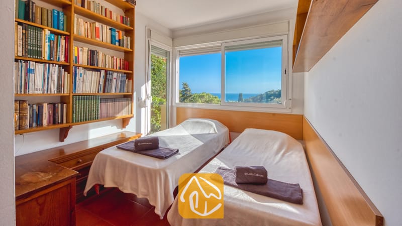 Villas de vacaciones Costa Brava España - Villa Santa Cristina - Dormitorio en Villa Santa Cristina con dos camas individuales, ropa de cama blanca y toallas grises. Una gran estantería de madera se encuentra junto a una ventana con vistas al mar.