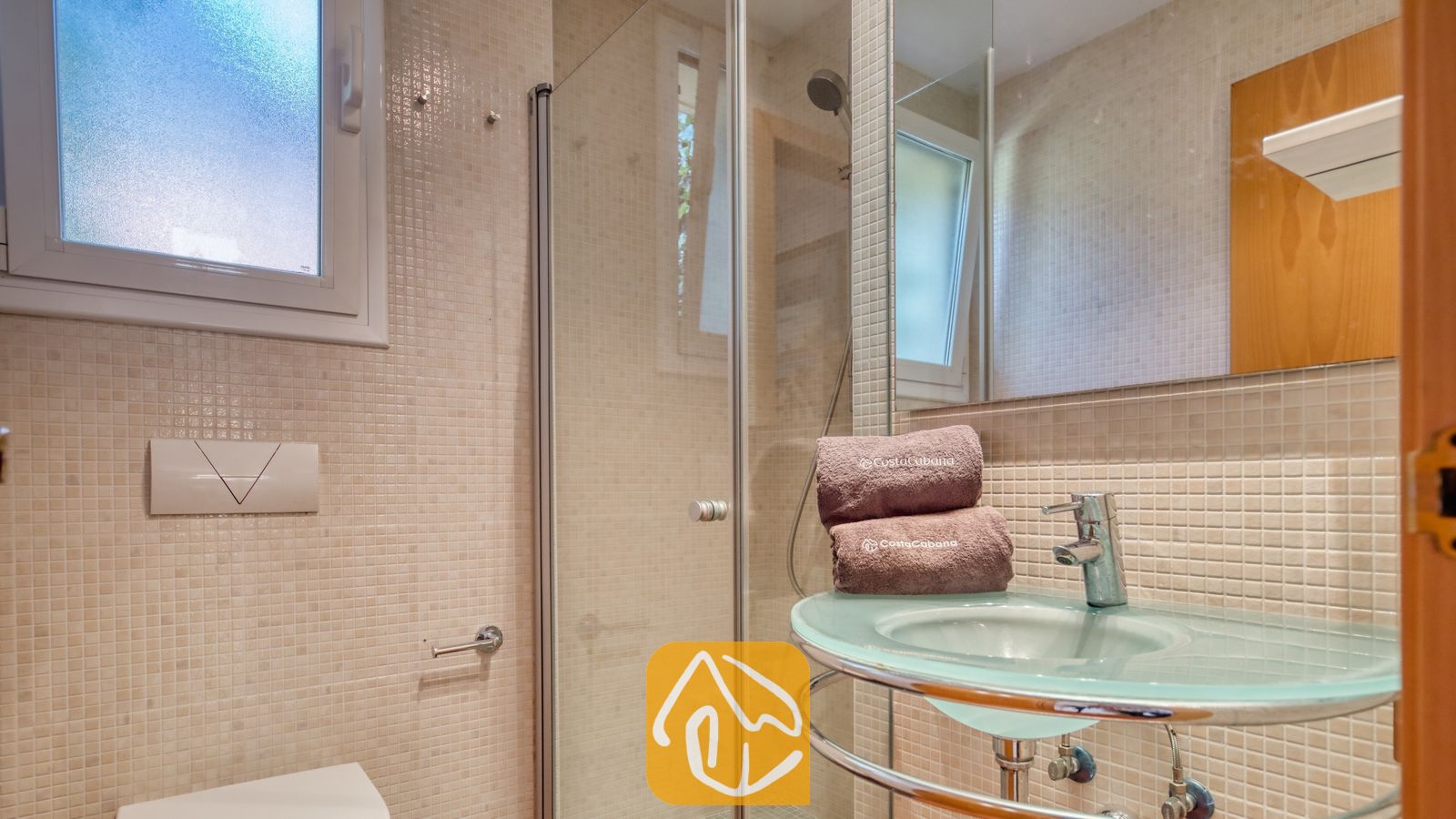 Vakantievilla's Costa Brava Spanje - Villa Santa Cristina - Badkamer met een glazen wastafel op zuil, chromen kraan en een inloopdouche. De wanden zijn betegeld met beige mozaïektegels en een grote spiegel reflecteert het matglazen raam.