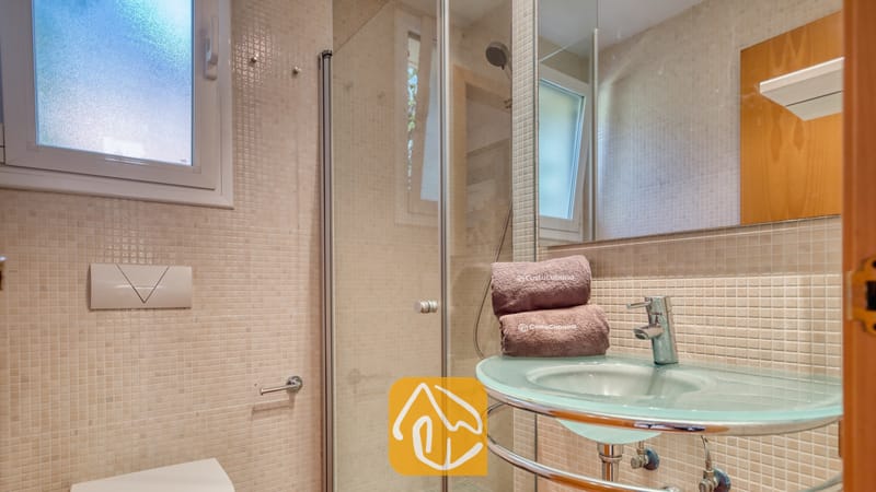 Villas de vacaciones Costa Brava España - Villa Santa Cristina - Baño con lavabo de pedestal de cristal, grifería cromada y ducha a ras de suelo. Las paredes están revestidas con azulejos de mosaico en tonos beige, y un gran espejo refleja una ventana esmerilada.