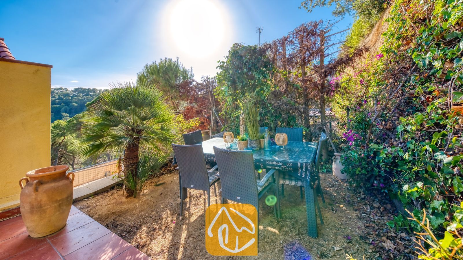 Vakantievilla's Costa Brava Spanje - Villa Santa Cristina - Buitenruimte om te dineren bij Villa Santa Cristina, voorzien van grijze rieten stoelen, een tafel met patroon en een kleine palmboom tegen een achtergrond van een weelderige, groene heuvel.