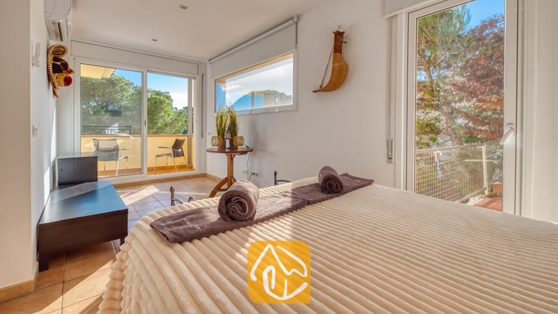 Vakantievilla's Costa Brava Spanje - Villa Santa Cristina - Slaapkamer met een crèmekleurige sprei met textuur en opgerolde paarse handdoeken, voorzien van grote glazen schuifdeuren die uitkomen op een zonnig terras bij Villa Santa Cristina.