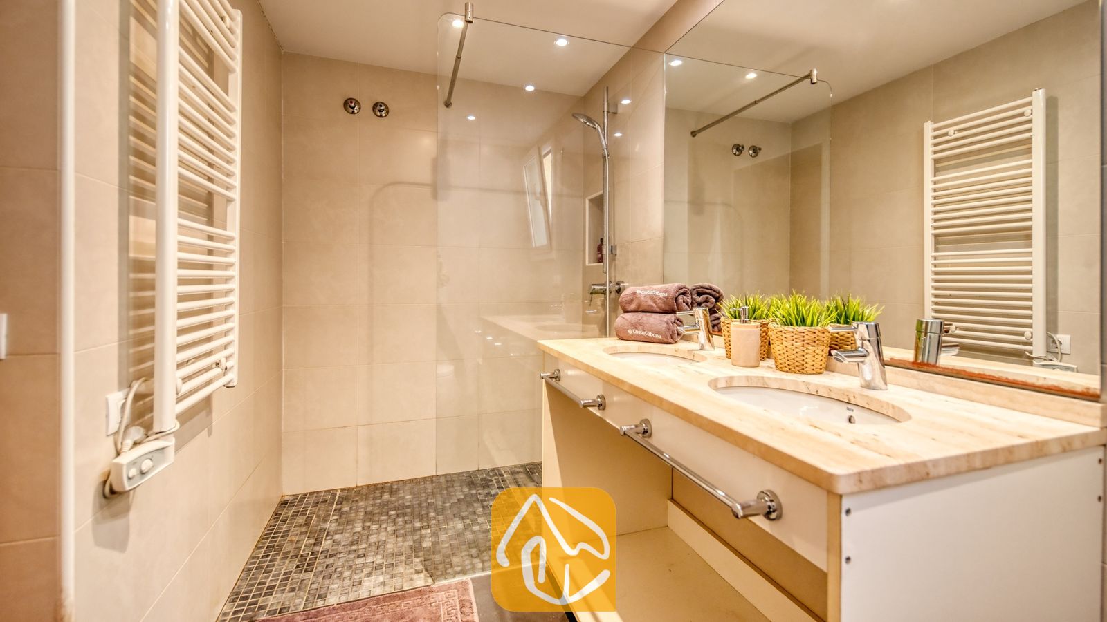 Vakantievilla's Costa Brava Spanje - Villa Santa Cristina - Badkamer voorzien van een dubbele wastafel, een grote spiegel en een inloopdouche met glazen wand en een vloer van donkere mozaïektegels.