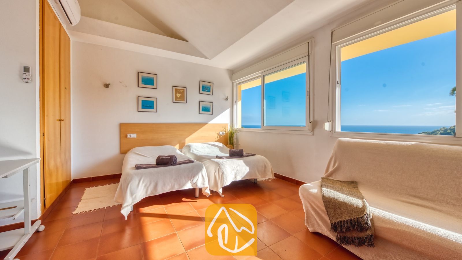 Vakantievilla's Costa Brava Spanje - Villa Santa Cristina - Lichte slaapkamer in Villa Santa Cristina, voorzien van twee eenpersoonsbedden, terracotta tegels en een witte bank. De grote ramen bieden een prachtig uitzicht op de blauwe oceaan onder een heldere hemel.