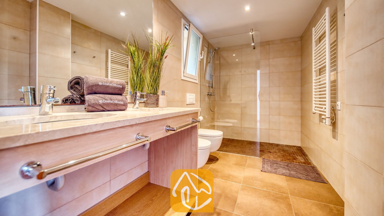 Vakantievilla's Costa Brava Spanje - Villa Santa Cristina - Moderne badkamer met dubbele wastafel, inloopdouche met glazen wand en beige betegelde muren. Opgerolde handdoeken en decoratieve planten sieren het stenen blad.