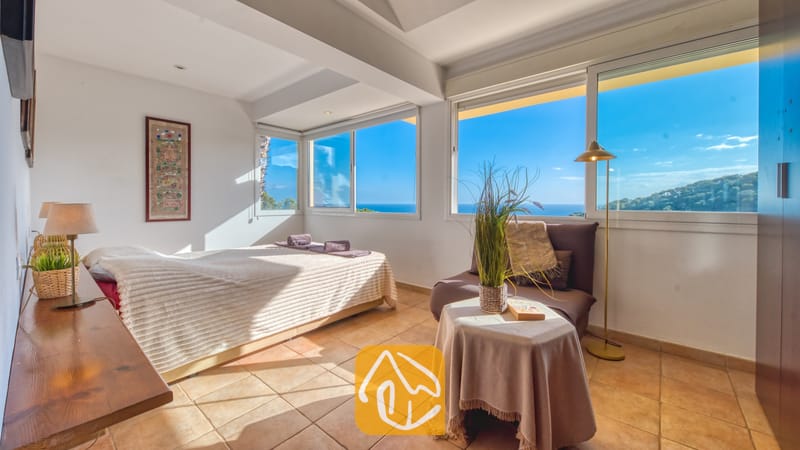 Vakantievilla's Costa Brava Spanje - Villa Santa Cristina - Slaapkamer in Villa Santa Cristina met een tweepersoonsbed, terracotta vloertegels en grote ramen die uitkijken over de zee en de heuvels langs de kust.
