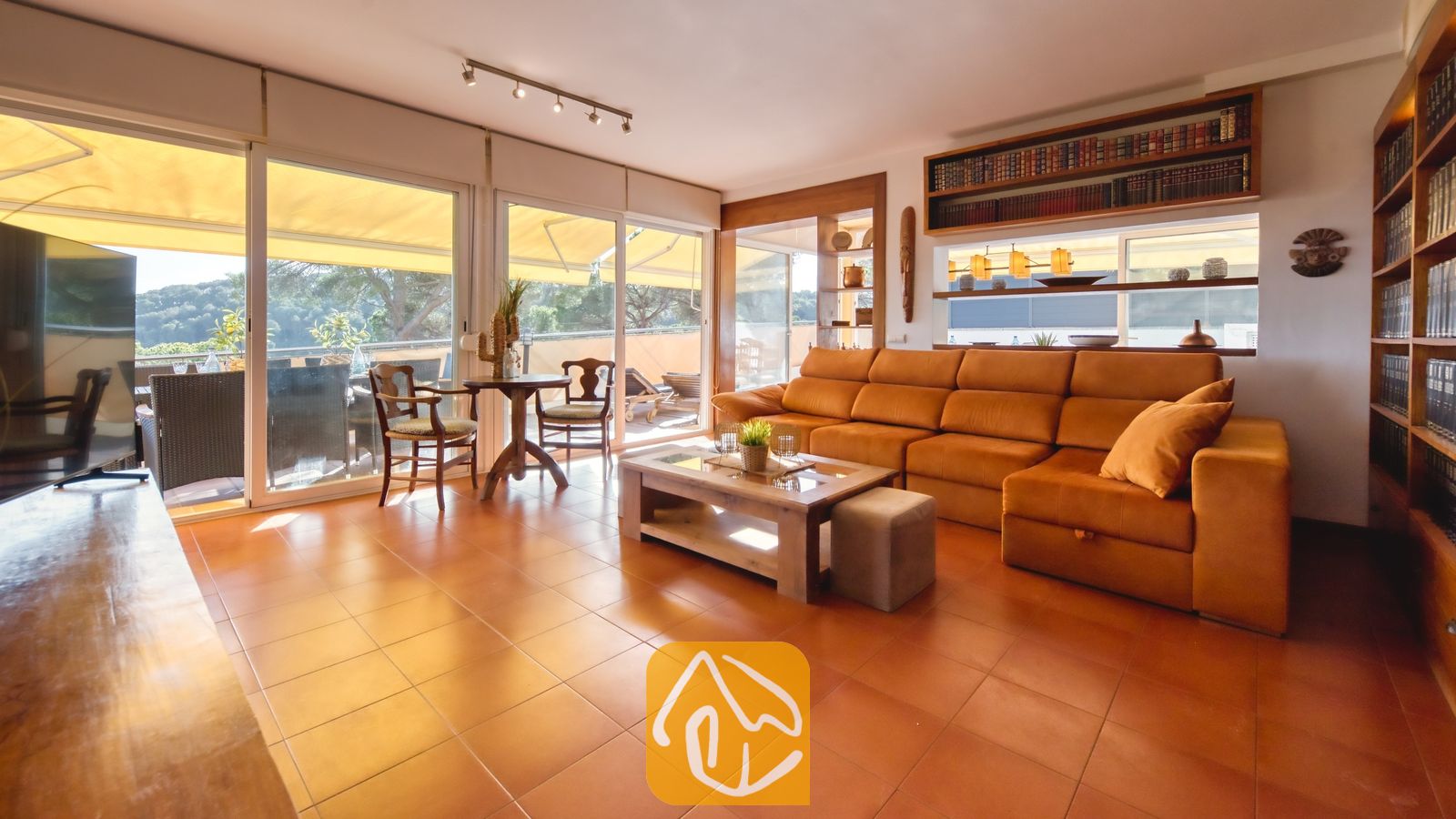 Vakantievilla's Costa Brava Spanje - Villa Santa Cristina - De woonkamer van Villa Santa Cristina is voorzien van een grote oranje hoekbank, terracotta plavuizen en ingebouwde houten boekenplanken naast de glazen schuifdeuren.