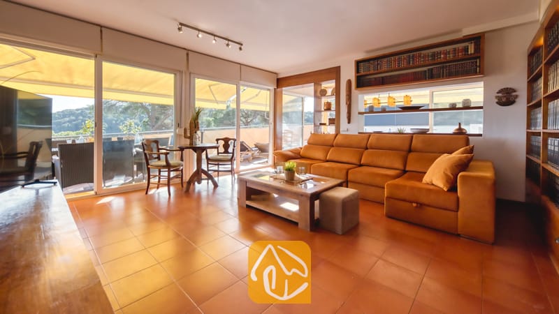 Vakantievilla's Costa Brava Spanje - Villa Santa Cristina - De woonkamer van Villa Santa Cristina is voorzien van een grote oranje hoekbank, terracotta plavuizen en ingebouwde houten boekenplanken naast de glazen schuifdeuren.
