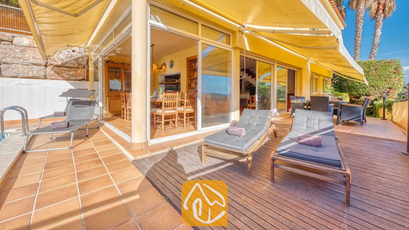 Vakantievilla's Costa Brava Spanje - Villa Santa Cristina - Terras van Villa Santa Cristina, voorzien van houten ligbedden met grijze kussens, grote gele zonneschermen en glazen deuren die toegang bieden tot een eethoek binnen.