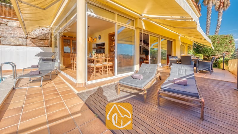 Vakantievilla's Costa Brava Spanje - Villa Santa Cristina - Terras van Villa Santa Cristina, voorzien van houten ligbedden met grijze kussens, grote gele zonneschermen en glazen deuren die toegang bieden tot een eethoek binnen.