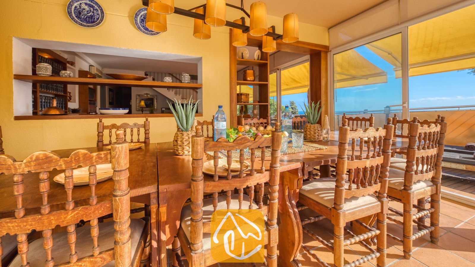 Vakantievilla's Costa Brava Spanje - Villa Santa Cristina - Grote houten eettafel met bewerkte stoelen en crèmekleurige kussens in Villa Santa Cristina. Gele muren, decoratieve blauwe borden en schuifdeuren die toegang bieden tot een zonovergoten terras.