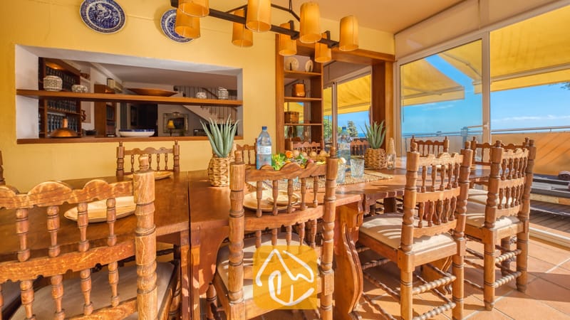 Vakantievilla's Costa Brava Spanje - Villa Santa Cristina - Grote houten eettafel met bewerkte stoelen en crèmekleurige kussens in Villa Santa Cristina. Gele muren, decoratieve blauwe borden en schuifdeuren die toegang bieden tot een zonovergoten terras.