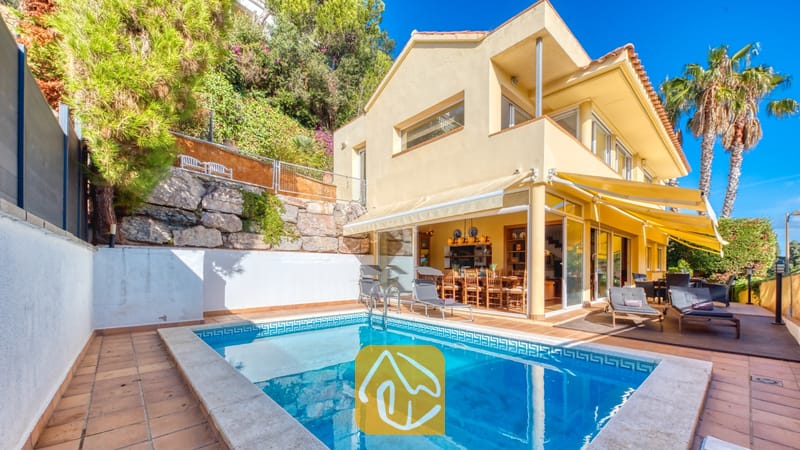 Vakantievilla's Costa Brava Spanje - Villa Santa Cristina - Privézwembad bij Villa Santa Cristina met een betegeld terras, gele zonneschermen en ligbedden op een houten vlonder naast de gele gevel.