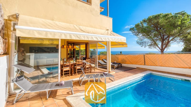 Vakantievilla's Costa Brava Spanje - Villa Santa Cristina - Zwembad en terras bij Villa Santa Cristina met terracotta tegels, grijze ligbedden en een houten eethoek in de schaduw van een geel zonnescherm, met uitzicht op zee.