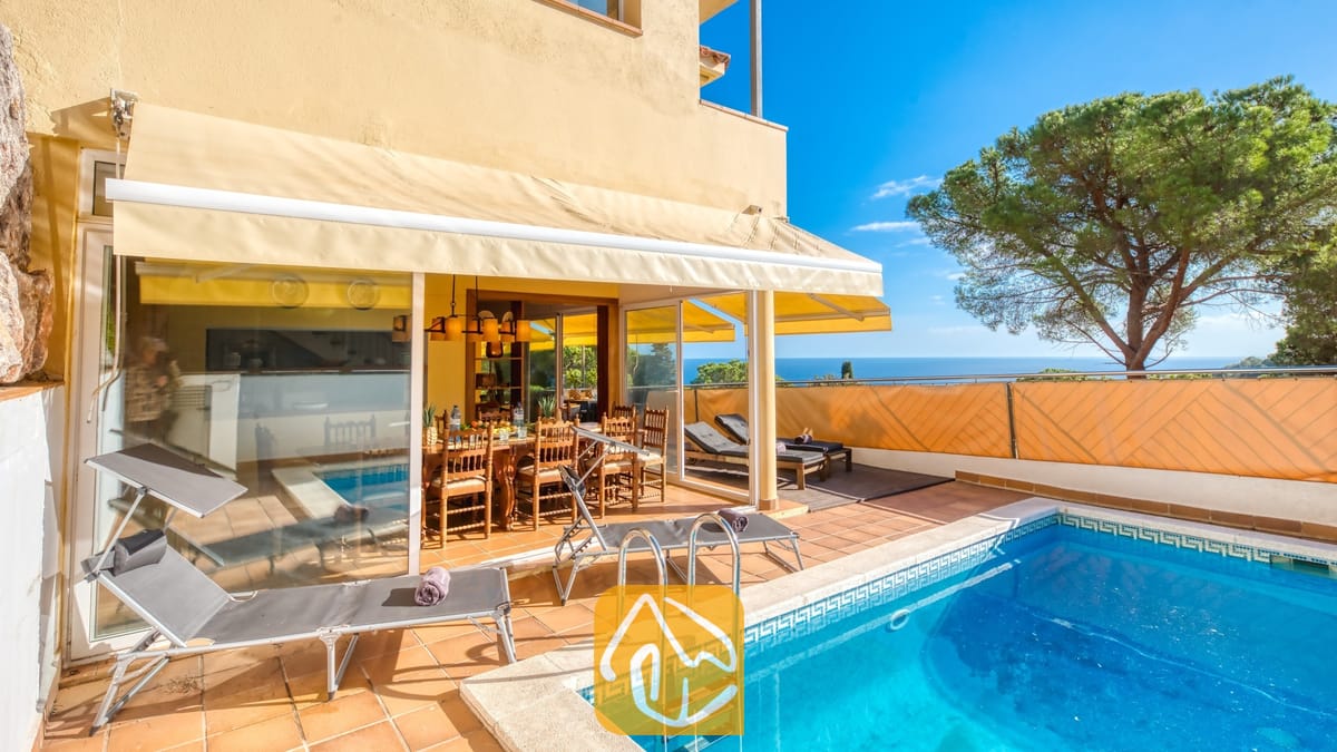 Vakantievilla's Costa Brava Spanje - Villa Santa Cristina - Zwembad en terras bij Villa Santa Cristina met terracotta tegels, grijze ligbedden en een houten eethoek in de schaduw van een geel zonnescherm, met uitzicht op zee.